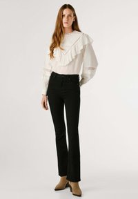 Blusa blanca con volantes y detalles de encaje, combinada con jeans acampanados negros y botas de tobillo beige, creando un look con capas y texturas.