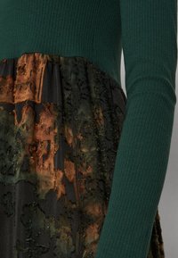 Manche en maille côtelée vert foncé et couture à la taille combinées avec une section de jupe en tissu texturé à fleurs noires et marron.