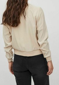 Beige bomber bunda s žebrovanými manžetami a lemem, s hladkou texturou. Nosená s tmavými, těsně padnoucími džíny. Viditelné švy na ramenou.