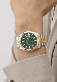 Reloj con esfera verde y pulsera de acero inoxidable en oro y plata; presenta marcadores de hora en negrita, display de fecha a las 3 en punto y bisel texturizado.