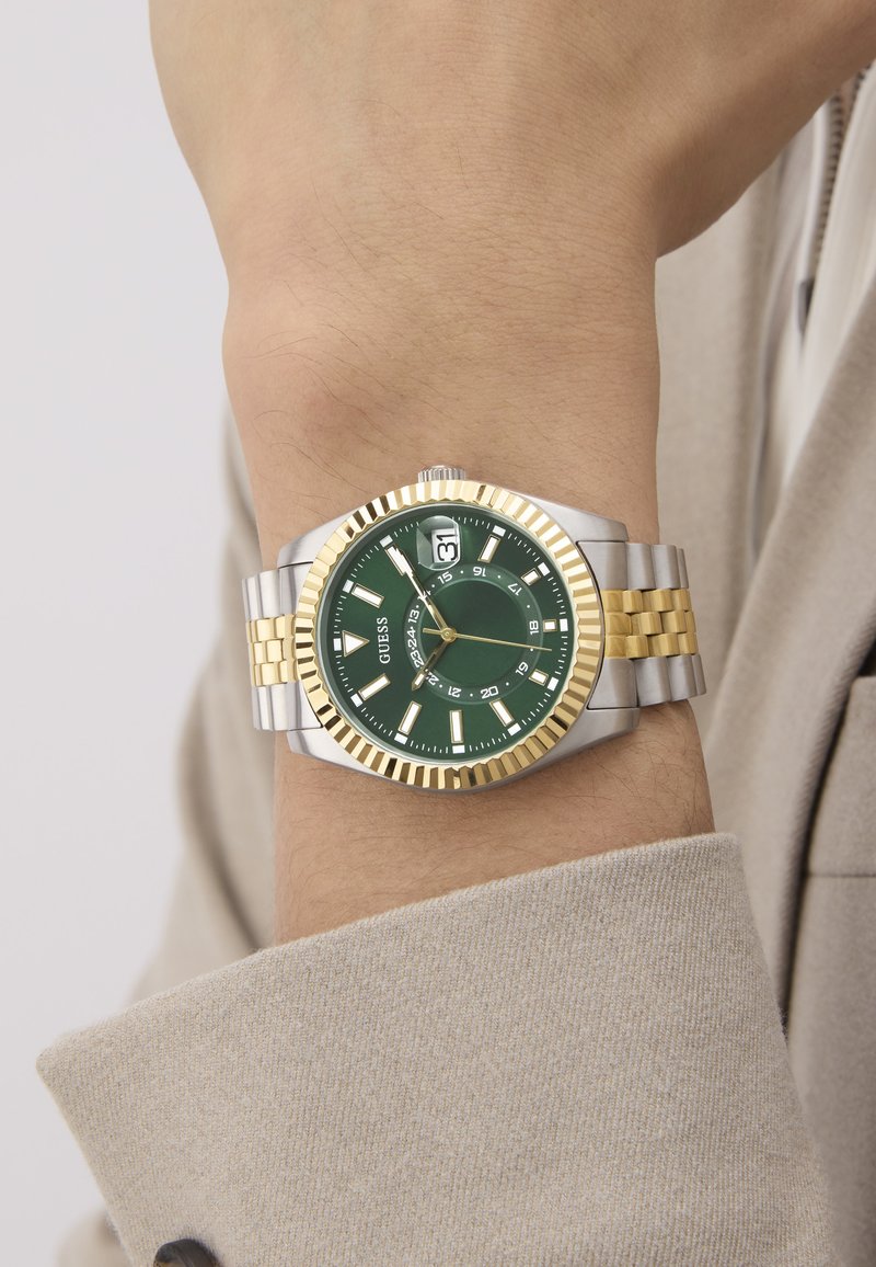 Reloj con esfera verde y pulsera de acero inoxidable en oro y plata; presenta marcadores de hora en negrita, display de fecha a las 3 en punto y bisel texturizado.