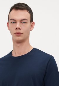 Tamsiai mėlynas medvilninis marškinėliai su apvaliu iškirpimu, trumpomis rankovėmis ir lygiu paviršiumi, prigludusio dizaino be raštų ar puošmenų.