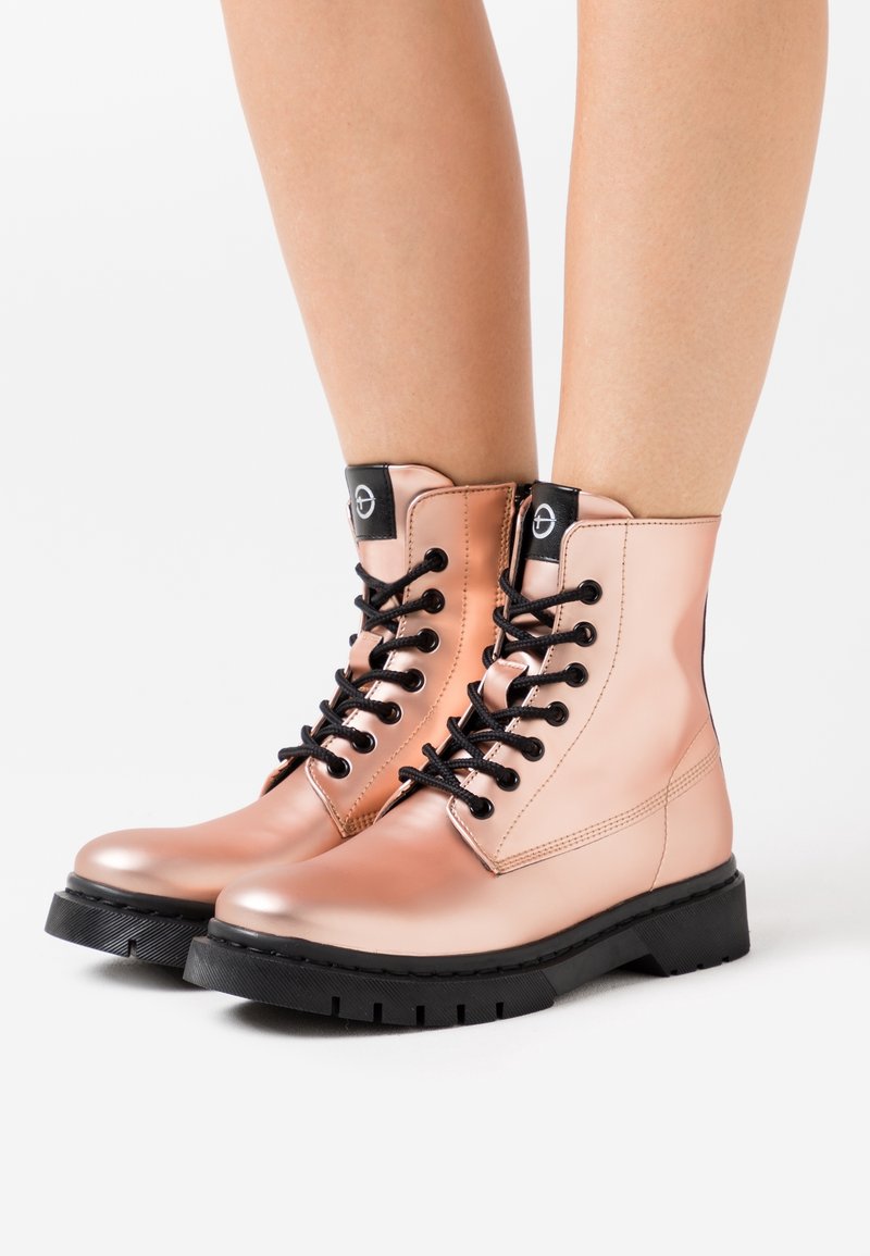 Jambes portant des bottines lacées rose clair brillantes avec semelles noires et œillets, debout sur un fond blanc uni.