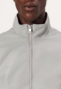 Lichtgrijze jas met een hoge kraag, volledige rits en een gladde, matte textuur. Minimalistisch ontwerp zonder zichtbare patronen.