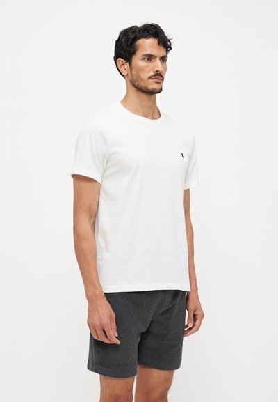 Tricou alb din bumbac cu guler rotund, mâneci scurte și un mic logo navy pe partea stângă a pieptului, purtat cu pantaloni scurți gri închis.