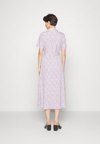 Vestido midi floral lilás com mangas curtas, gola e detalhe de cintura atada; feito de tecido suave, com um caimento solto. Sandálias pretas com salto bloco.
