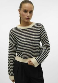 Gestreepte trui van breiwerk in crème en marineblauw met een textuurpatroon, ronde halslijn en geribbelde manchetten. Draagt met zwarte broek.