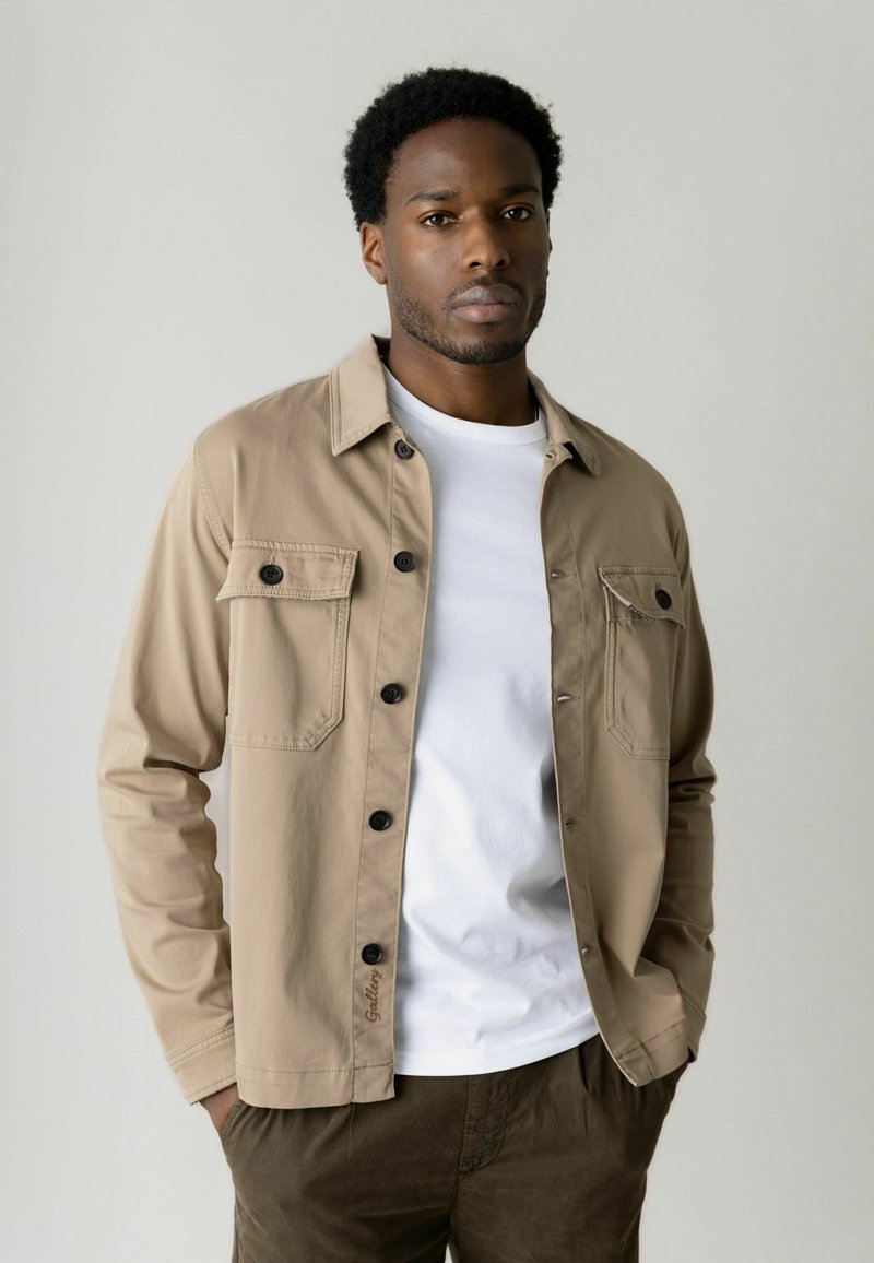 Lühikeste lokkis juustega mees kannab beeži nööbitud särki valge T-särgi kohal ja pruune pükse, seisab käed taskutes ühevärvilise tausta ees.