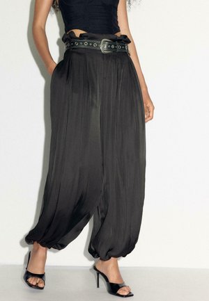 Kobieta nosząca luźne czarne satynowe spodnie haremowe z marszczonymi mankietami, czarny paskiem z ćwiekami, czarny krótki top i czarne wysokie buty na obcasie z odkrytymi palcami.