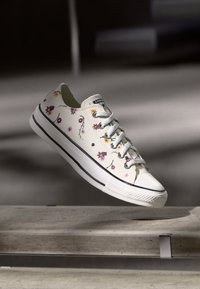 White Converse sneaker with floral print, white laces, and black stripe on sole, suspended above a concrete surface in soft lighting.
