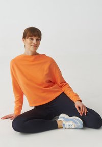Felpa arancione con una vestibilità rilassata e scollo ampio, abbinata a leggings blu navy e sneakers blu, su uno sfondo chiaro.