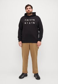 Čierna mikina s kapucňou a bielym nápisom "CALVIN KLEIN". Khaki nohavice, čierne tenisky s fialovými detailmi. Jednoduchý, casual dizajn.