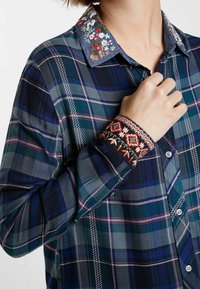 Persona che indossa una camicia di flanella a quadri blu con colletto floreale ricamato e polsini ricamati multicolori, mentre tiene la camicia vicino al colletto.