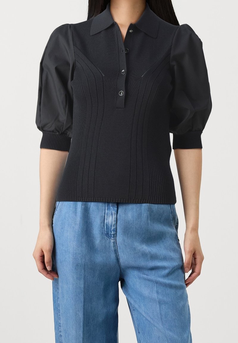 Zwarte gebreide polo met puffmouwen, geribbelde textuur, vijfknopenlijst en gestructureerde verticale lijnen. Gecombineerd met lichtblauwe denimbroek.