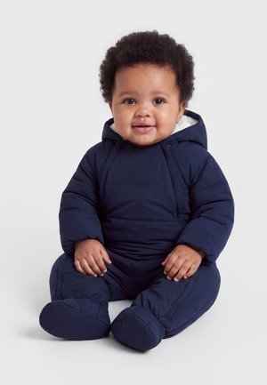Bébé souriant aux cheveux bouclés assis, portant une combinaison de ski matelassée bleu marine à capuche avec chaussons intégrés.