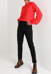 Punainen neulottu pusero, jossa on ilmapallohihat, yhdistetty korkeavyötäröisiin mustiin farkkuihin ja ruskeisiin, paksupohjaisiin nilkkureihin, joissa on nauhat.