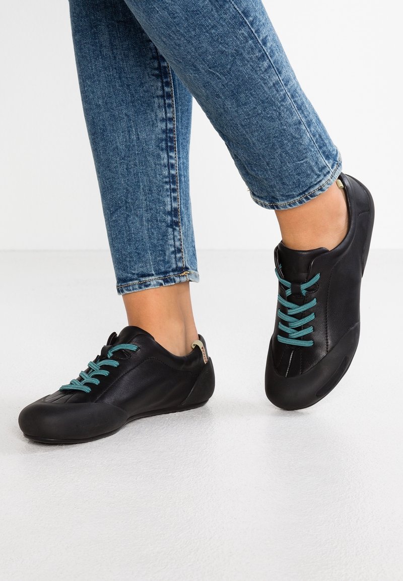 Personne portant des baskets en cuir noir avec des lacets turquoise et un jean en denim bleu, debout sur un sol blanc.