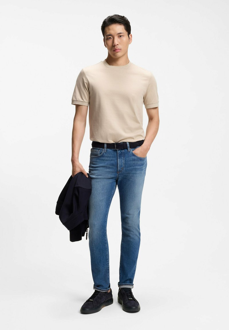 Jeune homme portant un t-shirt beige, un jean bleu, une ceinture noire et des chaussures, tenant une veste sombre dans une main, debout devant un fond blanc.