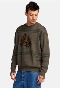 Bruine trui met een paardenprint, groene strepen en geribde manchetten. Voorzien van een ronde halslijn en logodetail in oranje aan de onderrand.
