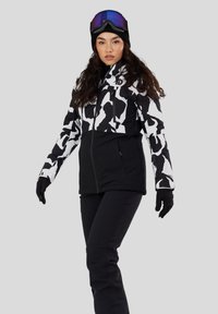 Frau trägt schwarz-weiß gemusterte Skijacke, schwarze Hose, schwarze Handschuhe, schwarze Mütze und Skibrille mit lila getönten Gläsern, steht vor grauem Hintergrund.