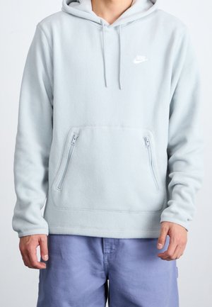 Person trägt hellgrauen Nike Kapuzenfleece mit vorderen Reißverschlusstaschen und blauer Shorts, steht vor einem schlichten weißen Hintergrund.