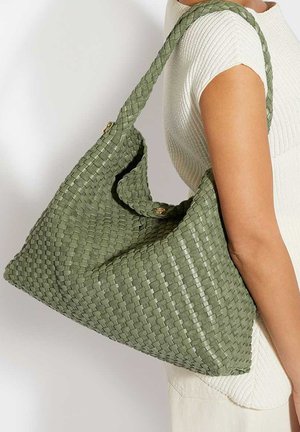 Frau trägt ein weißes Kurzarm-Top und eine große, gewebte olivgrüne Schultertasche mit breitem geflochtenem Riemen.