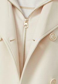 Beiger Mantel mit einem teilweise geschlossenen goldenen Reißverschluss, auf der Vorderseite sind Knöpfe mit der Gravur „S.Oliver“ zu sehen.