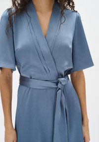Blaues, strukturiertes Wickelkleid mit kurzen Ärmeln, das eine taillierte Taille und einen gebundenen Gürtel hat. Glattes Material und dezentes Muster über das gesamte Kleid.