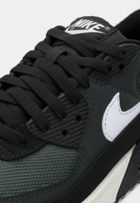 Nike-sneakers med överdel i mörkgrå mesh, svarta overlay, vitt logotyp och kontrasterande crème-sula. Funktioner med texturerade accenter och snörningsdesign.