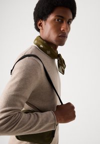 Mann in einem beigen Pullover und einem grün gemusterten Schal, der eine dunkelgrüne, strukturierte Umhängetasche mit einem schwarzen Gurt hält.