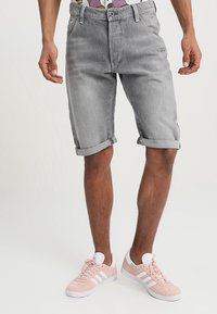 Hombre con pantalones cortos de mezclilla grises arremangados y zapatillas Adidas rosas con rayas blancas, de pie contra un fondo sencillo.