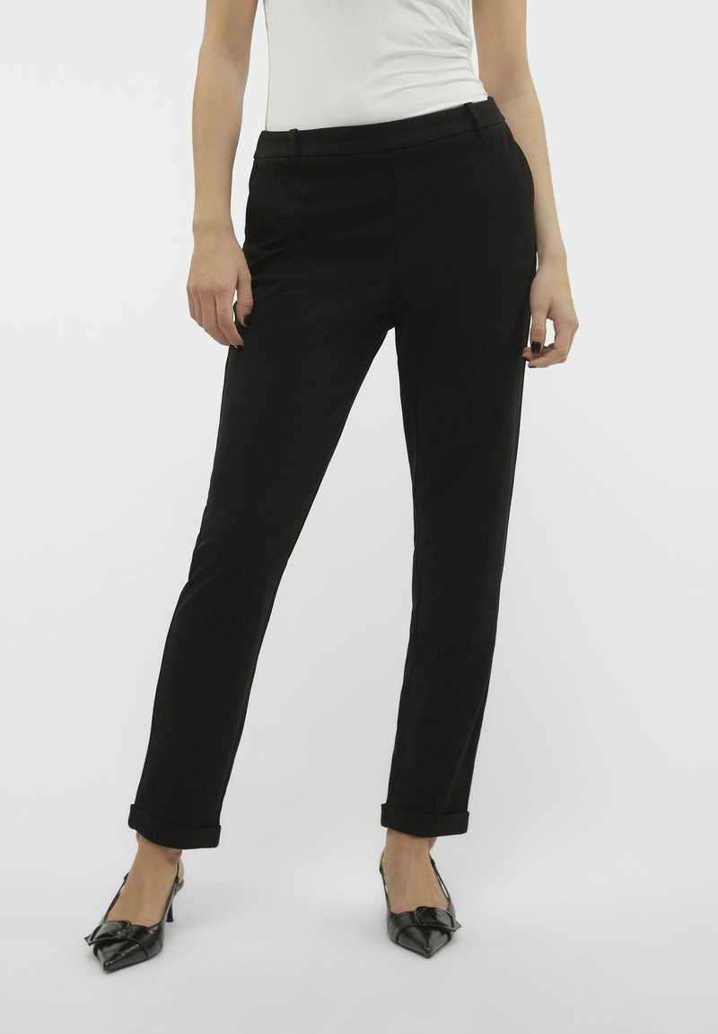 Zwarte broek van een gladde stof, met een getailleerde pasvorm, zijzakken en opgerolde mouwen; gecombineerd met puntige zwarte hakken.