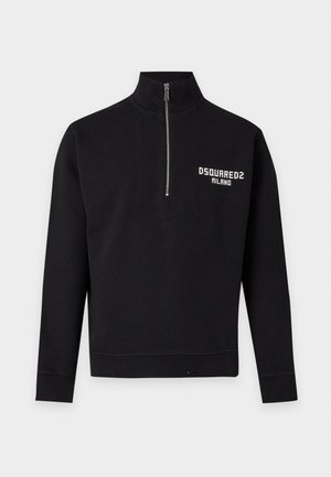 Jeune homme debout portant un sweat zippé noir avec le logo Dsquared2 Milano, un pantalon noir ample et des chaussures rouges sur un fond blanc uni.