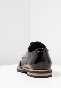 Chaussure en cuir verni noir avec une finition lisse, dotée d'une semelle en liège, d'un petit talon et d'un design à lacets. Lignes courbes et accents minimaux présents.