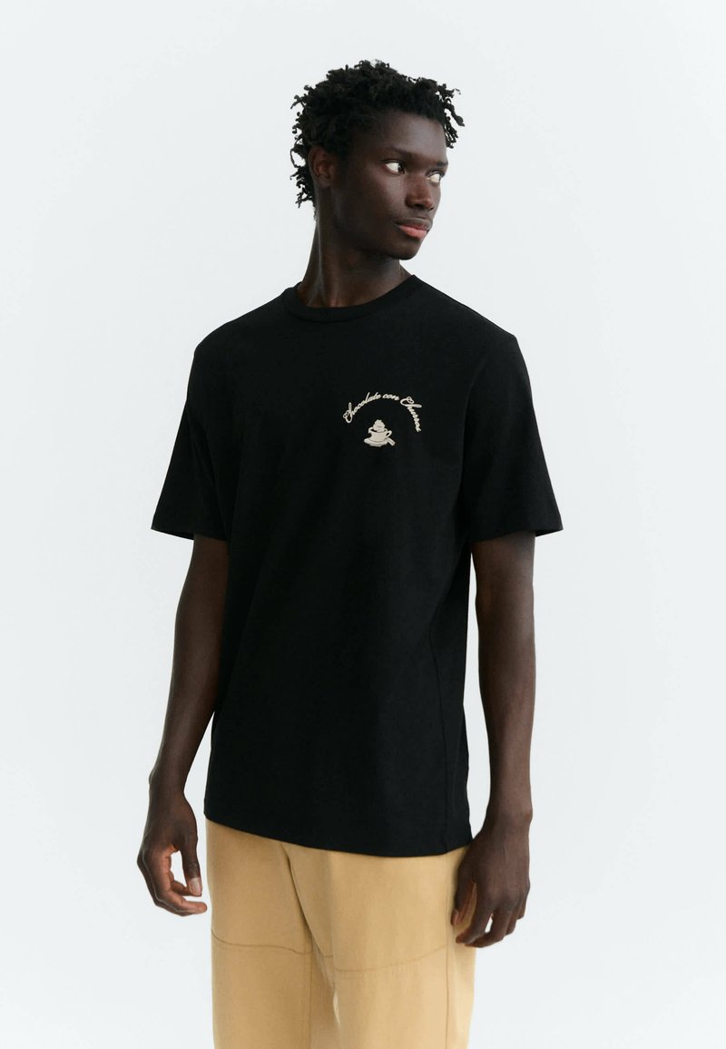 Jeune homme portant un t-shirt noir avec un texte et un graphisme beige, regardant sur le côté, associé à un pantalon beige clair sur un fond uni.