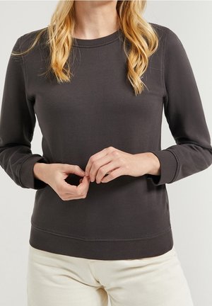 Blond osoba ubrana w ciemną koszulę z długim rękawem i jasne spodnie, z delikatnie splecionymi dłońmi na talii.