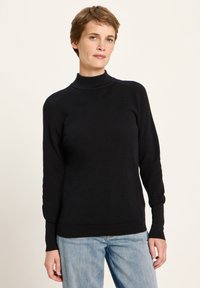 Frau mit kurzen braunen Haaren, die einen schwarzen Rollkragenpullover und hellblaue Jeans trägt und vor einem hellen, einfarbigen Hintergrund steht.