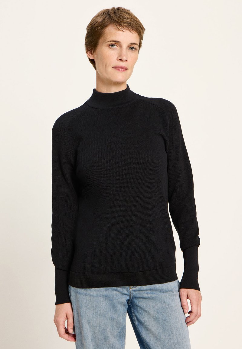 Frau mit kurzen braunen Haaren, die einen schwarzen Rollkragenpullover und hellblaue Jeans trägt und vor einem hellen, einfarbigen Hintergrund steht.