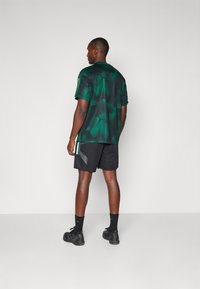 Green camo athletic shirt with a mesh texture, side stripes, paired with black shorts. Model stands from the back, showcasing the outfit.