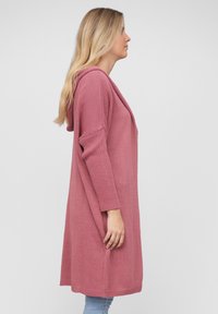 Lang rosa strikket cardigan med hette, åpen front og ribbestrikkede mansjetter. Har en avslappet passform og en midtlengde design.