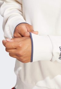 White sweatshirt with ribbed cuffs featuring blue accents. Image focuses on hands adjusting sleeve, highlighting texture and fit.