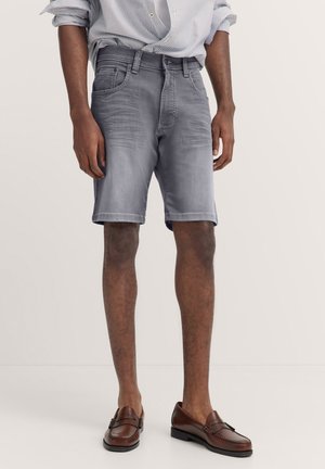 Graue Denim-Shorts mit klassischem Schnitt, ausgestattet mit Fronttaschen und einer leicht ausgebleichten Textur. Kombiniert mit braunen Slippers.