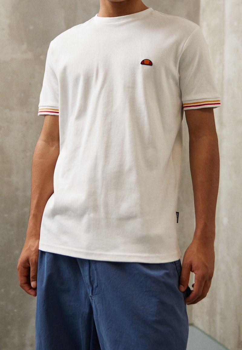 Homme portant un t-shirt blanc à manches courtes avec des poignets rayés rouges et jaunes, et un short bleu, debout contre un mur en béton gris.