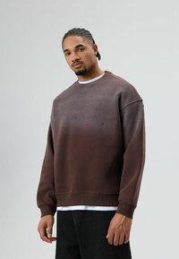 Bruin sweatshirt met verloopkleur, voorzien van gestippelde details, geribde manchetten en zoom. Gecombineerd met zwarte broek, gedragen door een model tegen een effen achtergrond.