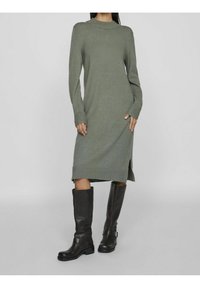 Knit dress in muted green with long sleeves, crew neck, side slits, paired with knee-high black leather boots. Soft texture, fitted design.
