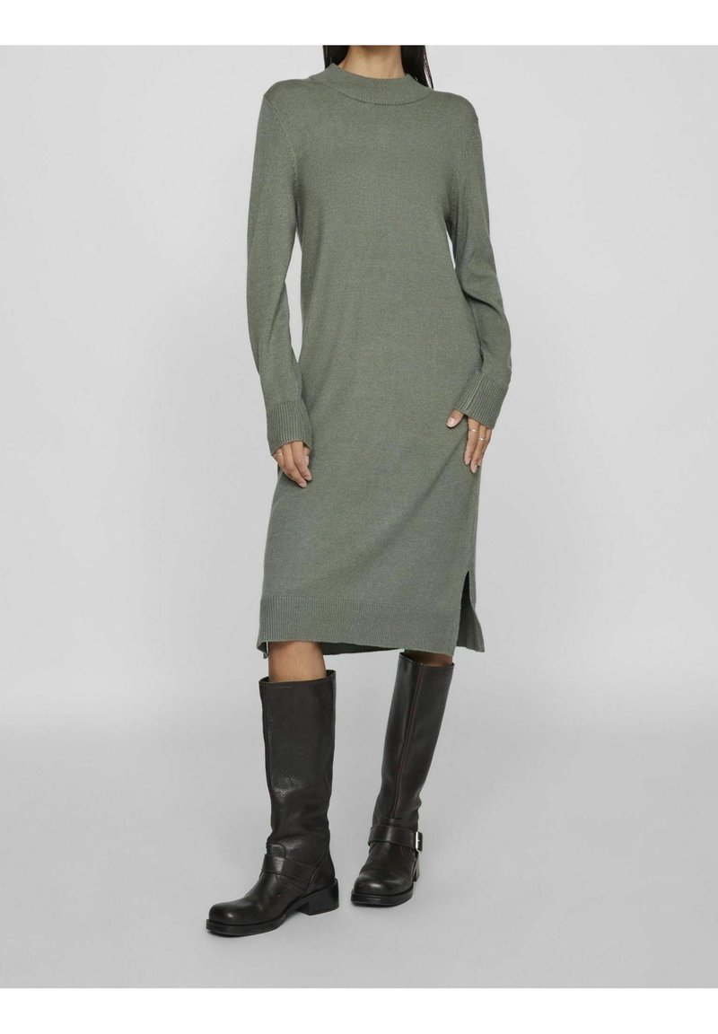 Knit dress in muted green with long sleeves, crew neck, side slits, paired with knee-high black leather boots. Soft texture, fitted design.