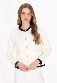 Cream cardigan with a round navy collar, gold button closures, and two front pockets. Features a soft texture and contrast trim.