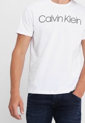 T-shirt blanc à manches courtes avec le texte "Calvin Klein", porté par une personne avec une main dans la poche d'un jean bleu foncé.