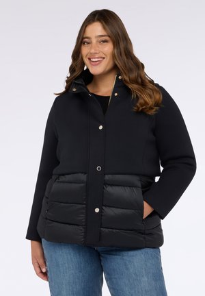 Schwarze Jacke mit einem glatten oberen Abschnitt und einem gepolsterten unteren Abschnitt. Mit Druckknöpfen und einem hohen Kragen. Über einem schwarzen Pullover getragen.