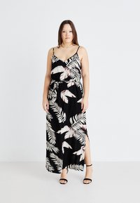 Robe maxi noire avec un motif de feuilles blanc et beige, décolleté en v, bretelles fines, taille marquée et fente latérale. Modèle portant des sandales noires à brides. 